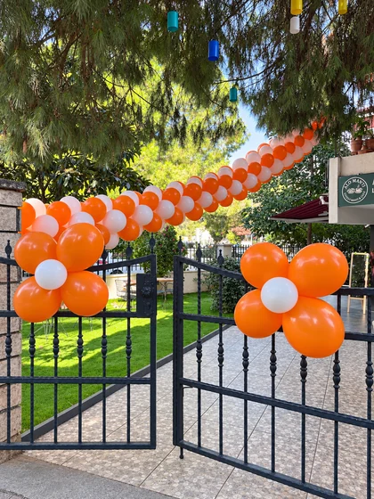 Siyah demir bahçe kapısında turuncu-beyaz balon zinciri ve iki çiçek biçimli balon süslemesi